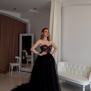 May include: A woman wearing a black strapless tulle gown with a lace bodice. The gown has a long train and is flowing. The woman is standing in a room with white walls and a white couch.