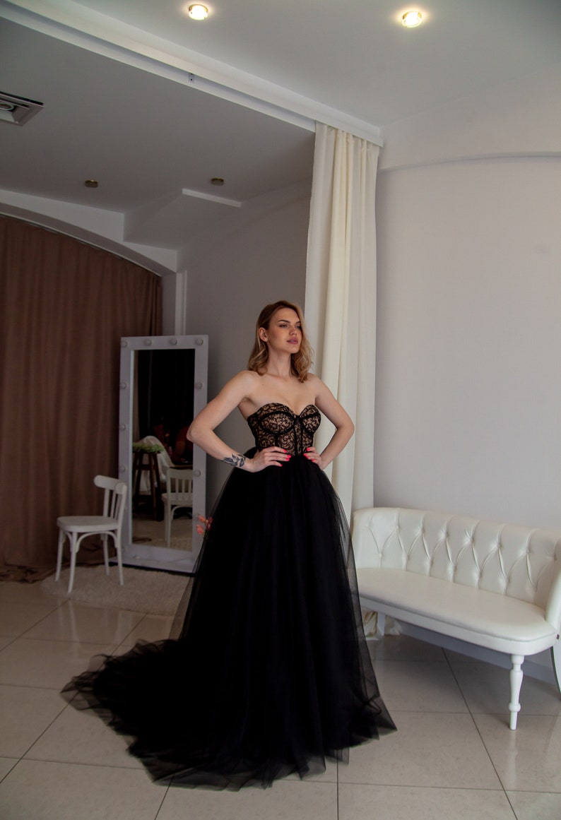May include: A woman wearing a black strapless tulle gown with a lace bodice. The gown has a long train and is flowing. The woman is standing in a room with white walls and a white couch.