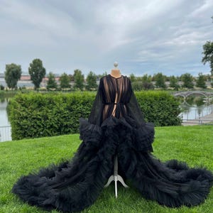 May include: A black tulle robe with long sleeves and a flowing train, displayed on a dress form outdoors. The robe features layers of ruffled fabric and a sheer design, creating a dramatic and elegant look. The background includes green grass, trees, and a cloudy sky.