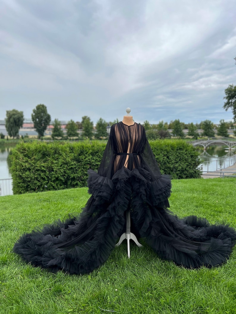 May include: A black tulle robe with long sleeves and a flowing train, displayed on a dress form outdoors. The robe features layers of ruffled fabric and a sheer design, creating a dramatic and elegant look. The background includes green grass, trees, and a cloudy sky.