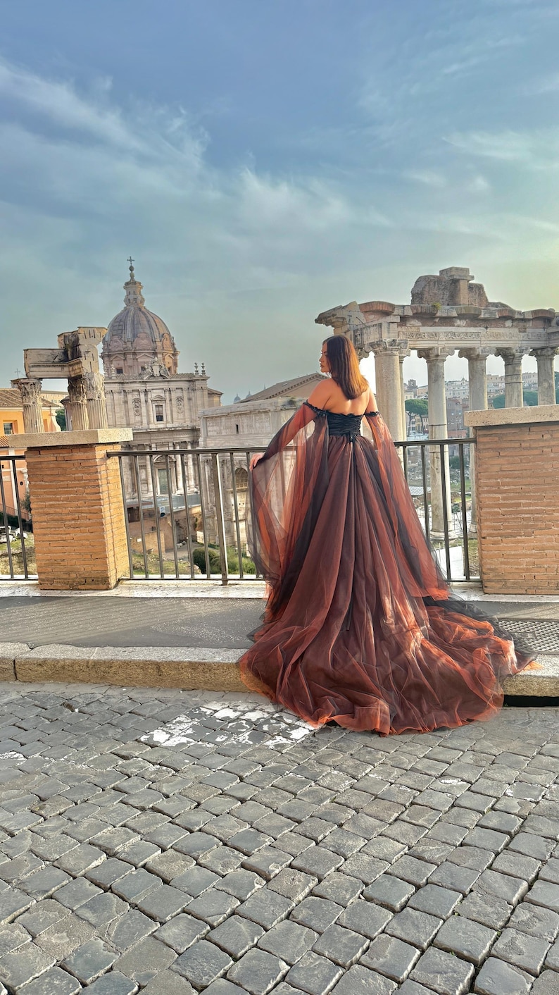 May include: A long, flowing gown with a black bodice and a layered, reddish-brown skirt. The dress features off-the-shoulder sleeves and a train. The background includes ancient Roman ruins and a cloudy sky, creating a dramatic and elegant aesthetic.