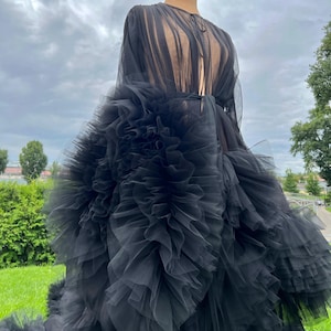 May include: A black tulle gown on a mannequin. The dress features a sheer, long-sleeved bodice and a voluminous skirt with layers of ruffled tulle. The gown is displayed outdoors against a backdrop of green grass and a cloudy sky.