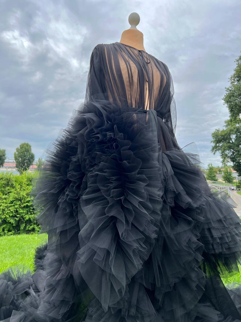 May include: A black tulle gown on a mannequin. The dress features a sheer, long-sleeved bodice and a voluminous skirt with layers of ruffled tulle. The gown is displayed outdoors against a backdrop of green grass and a cloudy sky.