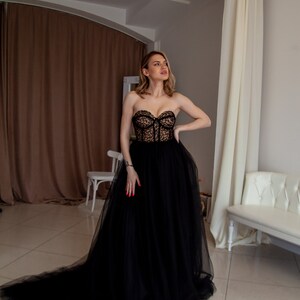 May include: A woman wearing a black strapless tulle gown with a lace bodice. The gown has a long train and the woman is standing in a room with white walls and a white couch.