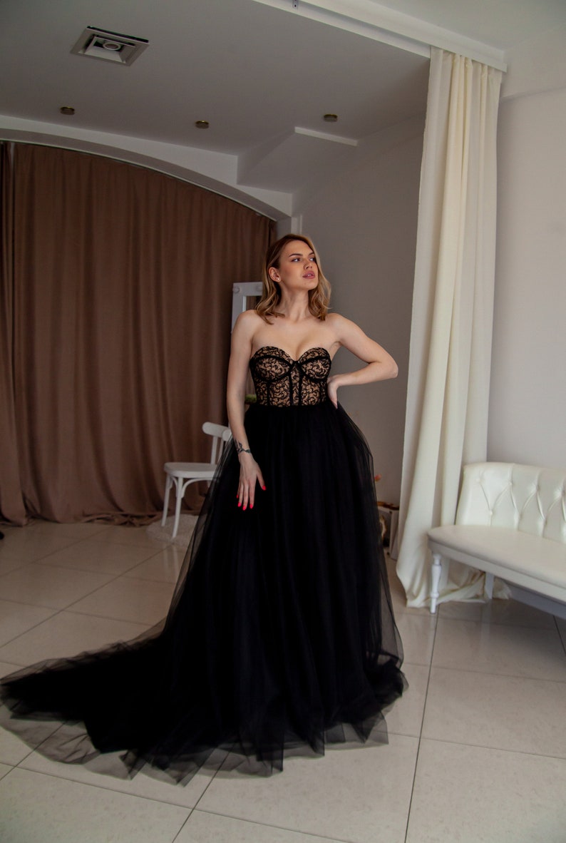 May include: A woman wearing a black strapless tulle gown with a lace bodice. The gown has a long train and the woman is standing in a room with white walls and a white couch.