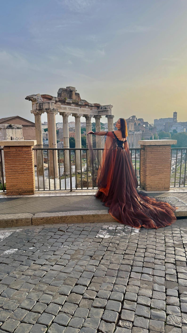 May include: A long, flowing gown with a dark brown skirt and a sheer, orange-toned cape. The dress features a black bodice and a long train. The background includes ancient Roman ruins and a cityscape.