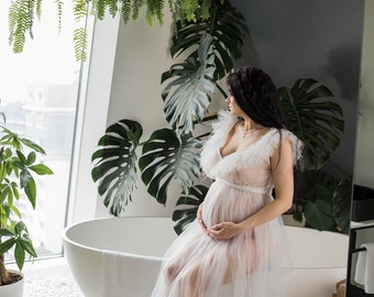 Könnte beinhalten: Eine schwangere Frau in einem weißen Tüllkleid sitzt am Rand einer weißen Badewanne. Sie ist umgeben von üppigen grünen Pflanzen, darunter Farne und Monstera-Blätter.