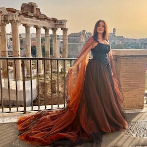 May include: A woman in a black and brown formal gown with a flowing train and cape stands on a balcony overlooking ancient ruins. The dress features a black bodice with intricate detailing and a layered skirt. The cape is a gradient of orange and brown.