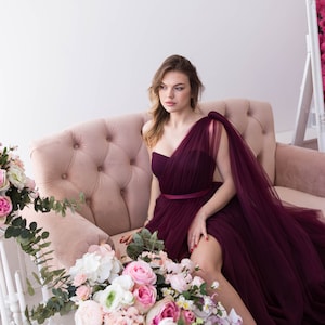 Puede incluir: Una mujer con un vestido de tul de un solo hombro de color burdeos se sienta en un sofá rosa. Lleva tacones altos y el pelo suelto. Hay flores delante de ella.