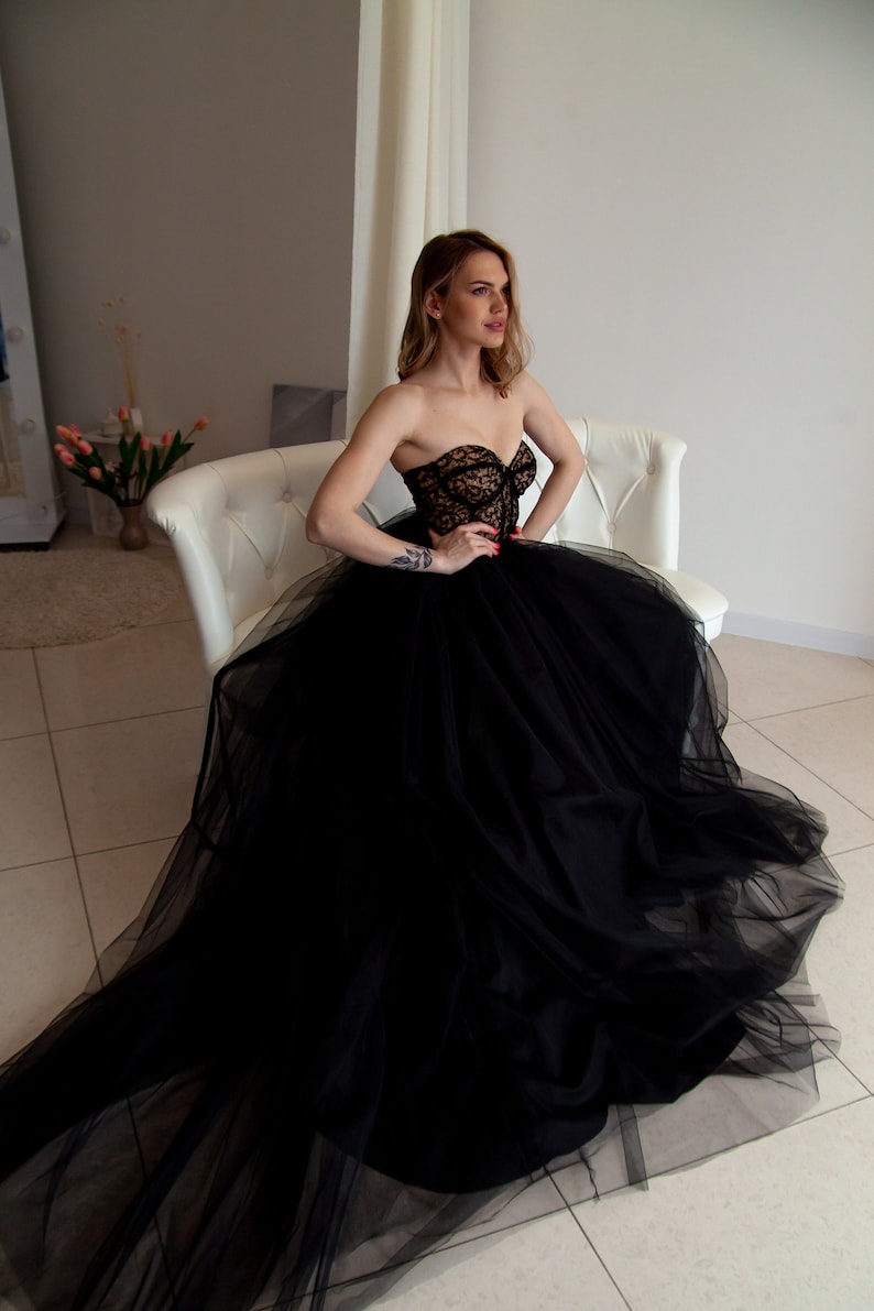May include: A woman wearing a black strapless tulle gown with lace detailing. She is sitting on a white couch with her hands on her lap.