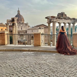 May include: A person wearing a brown off-the-shoulder gown with a long train, standing in front of ancient Roman ruins. The dress has a black bodice and a flowing, sheer skirt. The background features a cobblestone street and historic architecture.