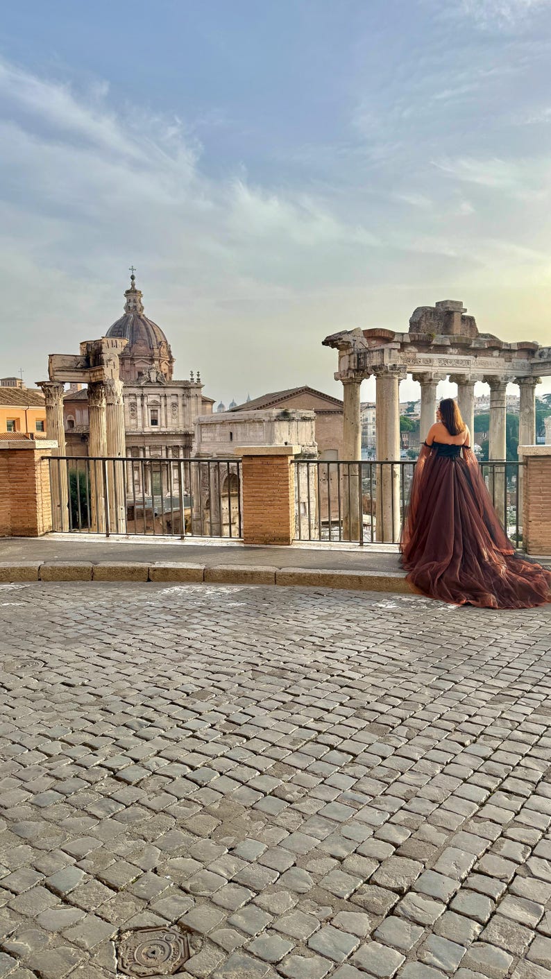 May include: A person wearing a brown off-the-shoulder gown with a long train, standing in front of ancient Roman ruins. The dress has a black bodice and a flowing, sheer skirt. The background features a cobblestone street and historic architecture.