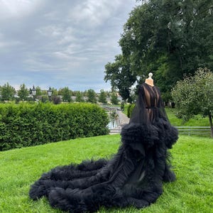 May include: A black tulle gown with a long train, displayed on a mannequin outdoors. The gown features a sheer bodice and ruffled detailing at the hem. The background includes green grass, trees, and a cloudy sky.