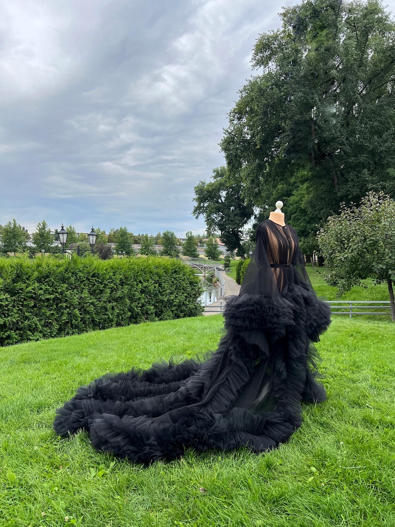 May include: A black tulle gown with a long train, displayed on a mannequin outdoors. The gown features a sheer bodice and ruffled detailing at the hem. The background includes green grass, trees, and a cloudy sky.