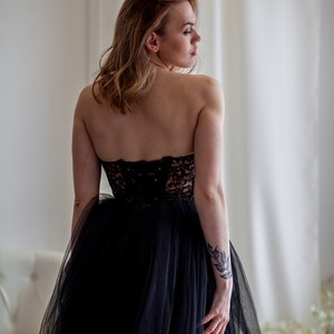 May include: A woman wearing a black strapless gown with a tulle skirt. She has a tattoo on her arm and is looking over her shoulder.
