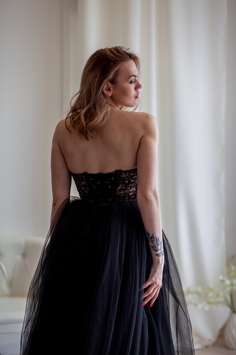 May include: A woman wearing a black strapless gown with a tulle skirt. She has a tattoo on her arm and is looking over her shoulder.