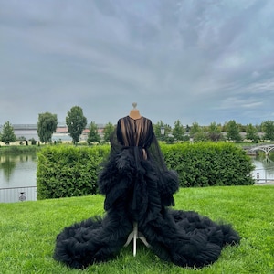May include: A black tulle robe with a long train is displayed on a mannequin in a grassy outdoor setting. The robe features a sheer bodice and a dramatic, ruffled skirt. The background includes green hedges, trees, and a cloudy sky.