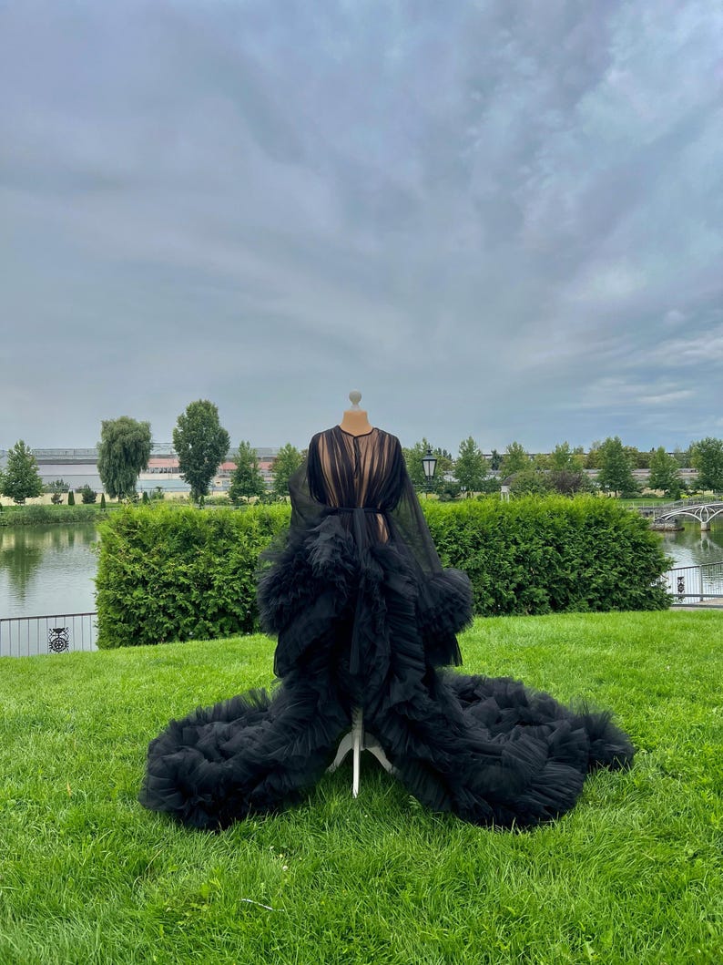 May include: A black tulle robe with a long train is displayed on a mannequin in a grassy outdoor setting. The robe features a sheer bodice and a dramatic, ruffled skirt. The background includes green hedges, trees, and a cloudy sky.