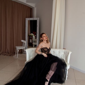 May include: A woman wearing a black strapless gown with a lace bodice and a tulle skirt sits on a white couch. The gown has a long train that flows behind her.