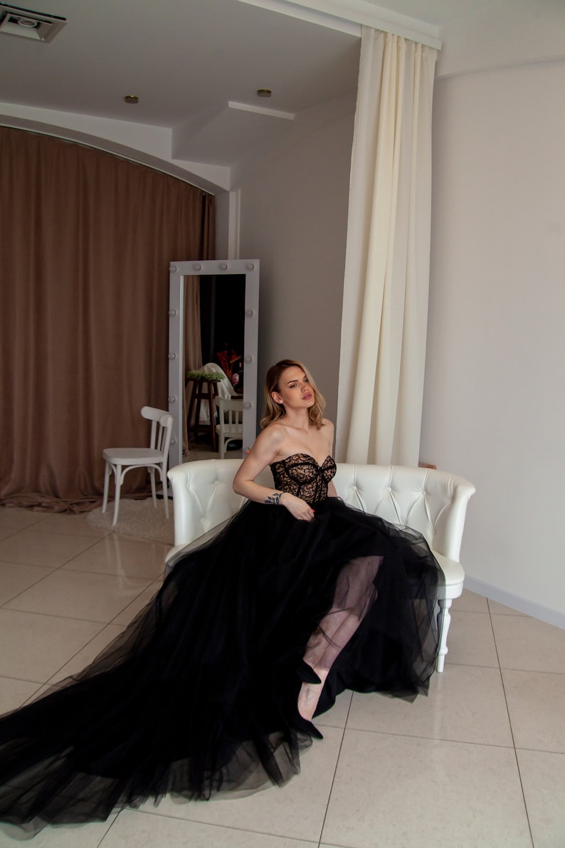 May include: A woman wearing a black strapless gown with a lace bodice and a tulle skirt sits on a white couch. The gown has a long train that flows behind her.
