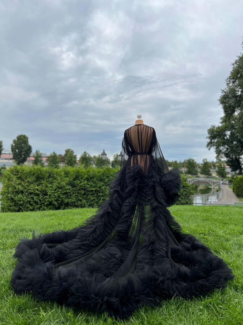 May include: A black tulle robe with a long train, displayed on a mannequin outdoors. The robe features a sheer bodice and sleeves, with a ruffled skirt that extends across the green grass. The background includes a cloudy sky and manicured landscaping.