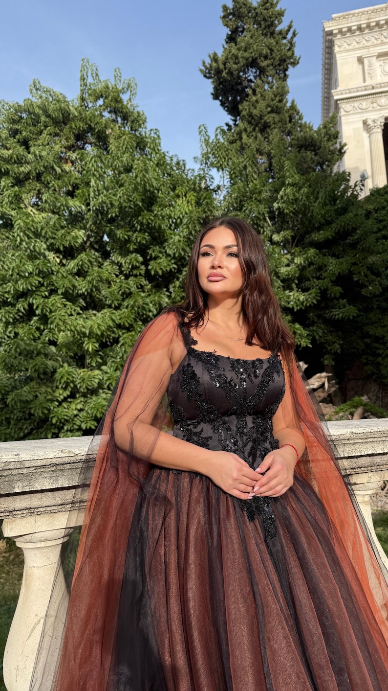 May include: A formal black gown with a tulle skirt and a sheer, rust-colored cape. The bodice is embellished with black lace and sequins. The dress is worn outdoors with a backdrop of greenery and a building.