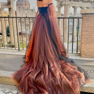 May include: A long, flowing gown with an off-the-shoulder design. The dress features a black bodice and a layered skirt in shades of brown and orange, with black accents. The dress is worn outdoors with a historical backdrop.