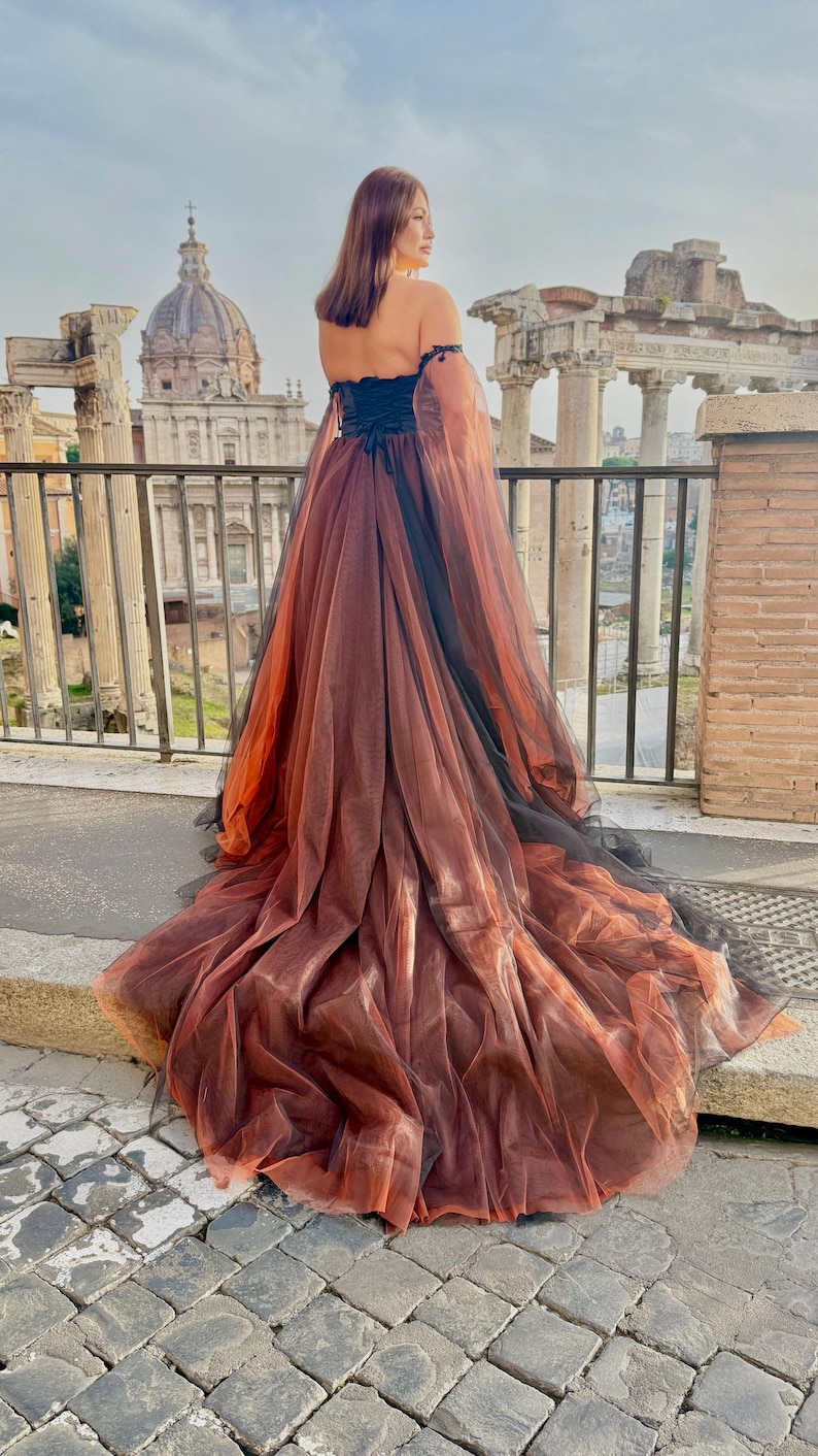 May include: A long, flowing gown with an off-the-shoulder design. The dress features a black bodice and a layered skirt in shades of brown and orange, with black accents. The dress is worn outdoors with a historical backdrop.