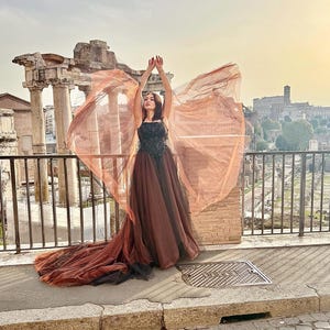 May include: A woman in a brown and black formal gown with a flowing train and sheer, wing-like fabric. The dress has a black bodice with intricate detailing. The background features ancient ruins and a cityscape under a bright sky.