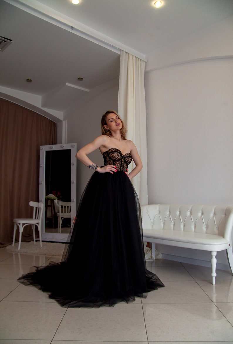 May include: A woman wearing a black strapless tulle gown with a lace bodice. The gown has a full skirt and is flowing. The woman is standing in a room with white walls and a white couch.