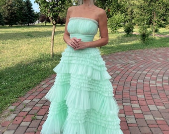 May include: A light green tulle dress with a strapless bodice and multiple layers of ruffles. The dress is worn by a woman standing on a brick pathway.
