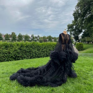 May include: A black tulle gown with a long train, displayed on a mannequin outdoors. The dress features a sheer bodice, long sleeves, and a ruffled skirt. The gown is set against a backdrop of green grass, trees, and a cloudy sky.