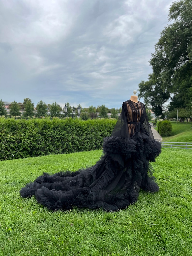 May include: A black tulle gown with a long train, displayed on a mannequin outdoors. The dress features a sheer bodice, long sleeves, and a ruffled skirt. The gown is set against a backdrop of green grass, trees, and a cloudy sky.