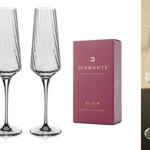 Op de afbeelding: Twee heldere glazen champagneflutes met een textuurontwerp. De flutes staan op een wit oppervlak. Een bordeauxrode doos met gouden letters luidt "Diamante Fine Lead Free Crystal Elixir Collection Beauty in its Element".