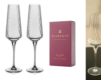 May include: Two clear glass champagne flutes with a textured design. The flutes are on a white surface. A burgundy box with gold lettering reads "Diamante Fine Lead Free Crystal Elixir Collection Beauty in its Element".