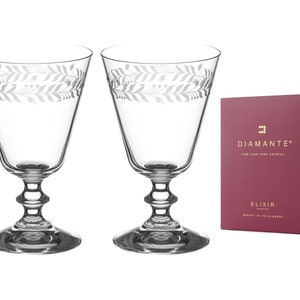 Puede incluir: Dos copas de vino de cristal transparente con un patrón decorativo de hojas alrededor del borde. Las copas tienen un tallo y una base detallados. Una caja granate con el texto "DIAMANTE" y "ELIXIR" está a la derecha de las copas.