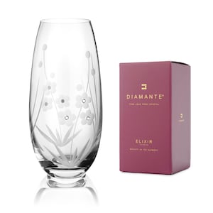 May include: A clear glass vase with a floral design etched into the side. The vase is sitting on a white surface and has a red box behind it. The box has the text "Diamante" and "Elixir" on it.