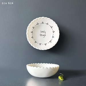 Könnte beinhalten: Zwei weiße Keramikschalen. Die obere Schale hat einen gewellten Rand und ein schwarzes Blumendesign am Rand. In der Mitte steht "lovely things". Die untere Schale ist ebenfalls weiß mit gewelltem Rand. Der Durchmesser der oberen Schale beträgt 6 cm.