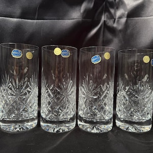 May include: Four clear, tall drinking glasses with intricate cut crystal designs. Each glass has a blue and gold sticker near the rim. The glasses are cylindrical with a slightly flared base, perfect for serving beverages.