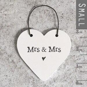Könnte beinhalten: Ein weißes herzförmiges Schild mit dem schwarzen Text „Mrs & Mrs“ und einem kleinen schwarzen Herz darunter. Das Schild ist etwa 3 cm hoch und hat eine schwarze Schnur zum Aufhängen. Der Hintergrund ist eine strukturierte graue Oberfläche.