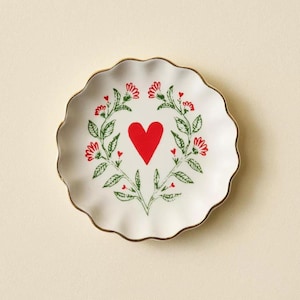 May include: A white ceramic plate with a scalloped edge and gold trim. The plate features a red heart in the center, surrounded by a green floral wreath with red flower accents. A decorative piece for the home.