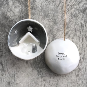 May include: Two white ceramic Christmas ornaments. One features a nativity scene inside a bowl with a gray and white background. The other is a white sphere with the text "Jesus, Mary and Joseph". Both are suspended by twine.