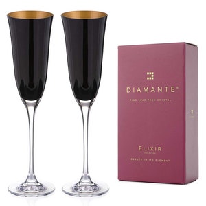 Könnte beinhalten: Zwei schwarze Sektgläser mit goldenen Rändern und klaren Stielen und Böden. Neben den Gläsern befindet sich eine bordeauxfarbene Schachtel mit goldenem Text und dem Wort "Diamante". Die Schachtel trägt auch die Worte "Elixir Collection" und "Beauty in its element".