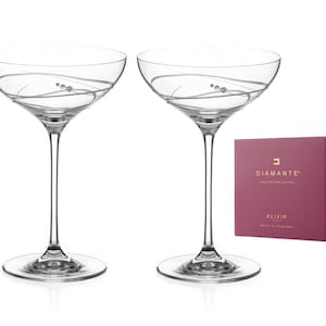Könnte beinhalten: Zwei klare Champagnergläser mit einem zarten Schwungmuster und einem einzelnen Kristall als Verzierung. Die Gläser stehen auf einem weißen Hintergrund mit einer rosafarbenen Schachtel, auf der "Diamante" und "Elixir" in goldenen Buchstaben stehen.