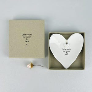 Può includere: Un piatto in ceramica bianco a forma di cuore con un piccolo ciondolo a forma di cuore, presentato in una scatola beige. Il piatto e la scatola presentano la scritta "Love you to the moon & back". Un piccolo fiore secco è accanto alla scatola.