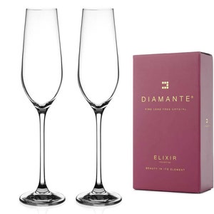Op de afbeelding: Twee heldere champagneglazen met lange stelen en ronde voetjes staan naast een bordeauxrode doos. De doos heeft gouden letters met de tekst "DIAMANTE" en "ELIXIR COLLECTION". Er staat ook "FINE LEAD FREE CRYSTAL" en "BEAUTY IN ITS ELEMENT".