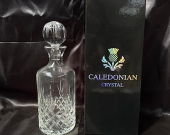 May include: A clear crystal decanter with a round stopper and diamond-cut pattern. Next to it is a black box with the words "CALEDONIAN CRYSTAL" and a thistle logo. The decanter is likely for spirits or wine.