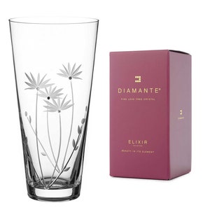 Puede incluir: Un jarrón de vidrio transparente con un diseño floral grabado en el lateral. El jarrón está sobre un fondo blanco y tiene una caja con el texto "Diamante Fine Lead Free Crystal Elixir Collection Beauty in its Element" en ella.