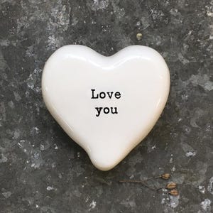Può includere: Un cuore in ceramica bianca con le parole "Love you" stampate in nero. Il cuore è liscio e lucido, appoggiato su una superficie grigia strutturata. Un piccolo rametto di fiori secchi è vicino al cuore.