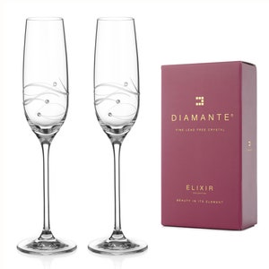 May include: Two clear glass champagne flutes with a delicate swirl design and small crystal accents. The flutes are on a white background with a burgundy box that says "Diamante Fine Lead Free Crystal Elixir Collection Beauty in its Element".